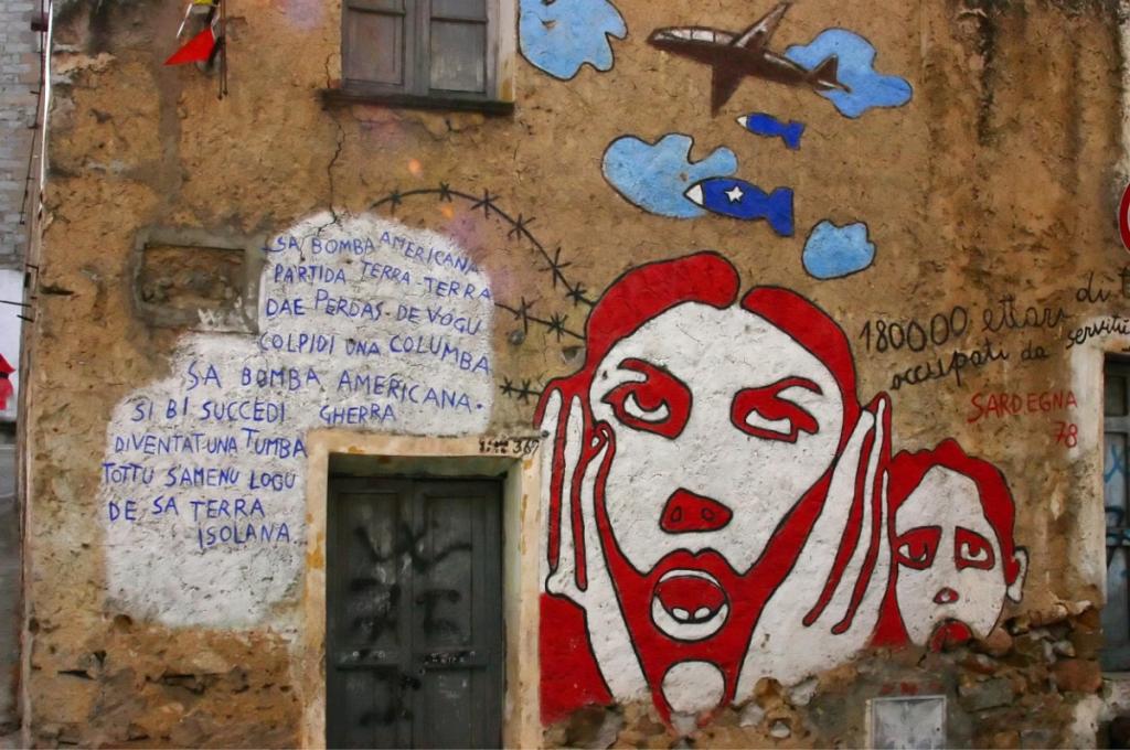 Murale con testo in sardo e immagini di guerra, aerei e volti stilizzati.