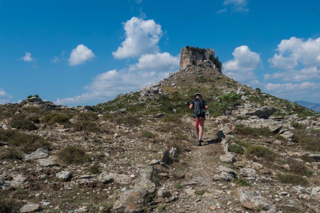 Escursionista cammina verso una cima rocciosa sotto un cielo azzurro con nuvole.