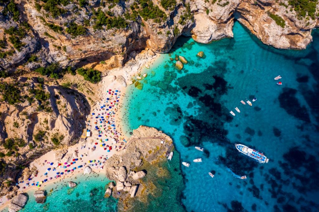 Spiaggia con acque cristalline, barche e ombrelloni colorati, circondata da scogliere.