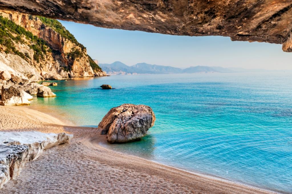 Spiaggia con sabbia dorata, rocce e mare cristallino sotto un cielo sereno.