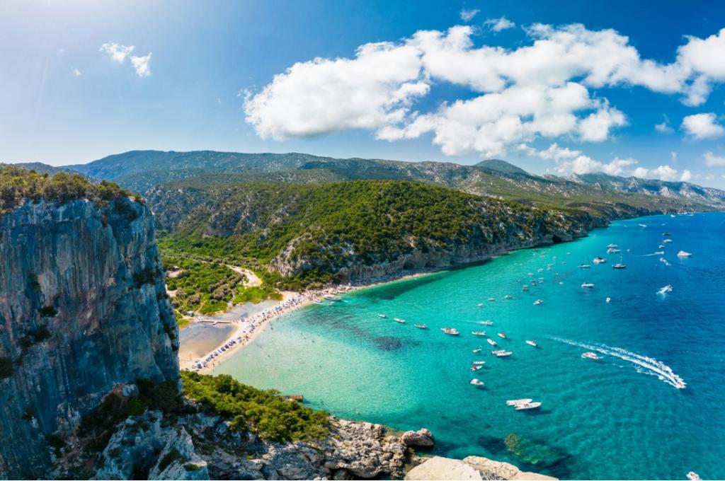 Spiaggia con acque turchesi, barche e scogliere verdi in un paesaggio mediterraneo.