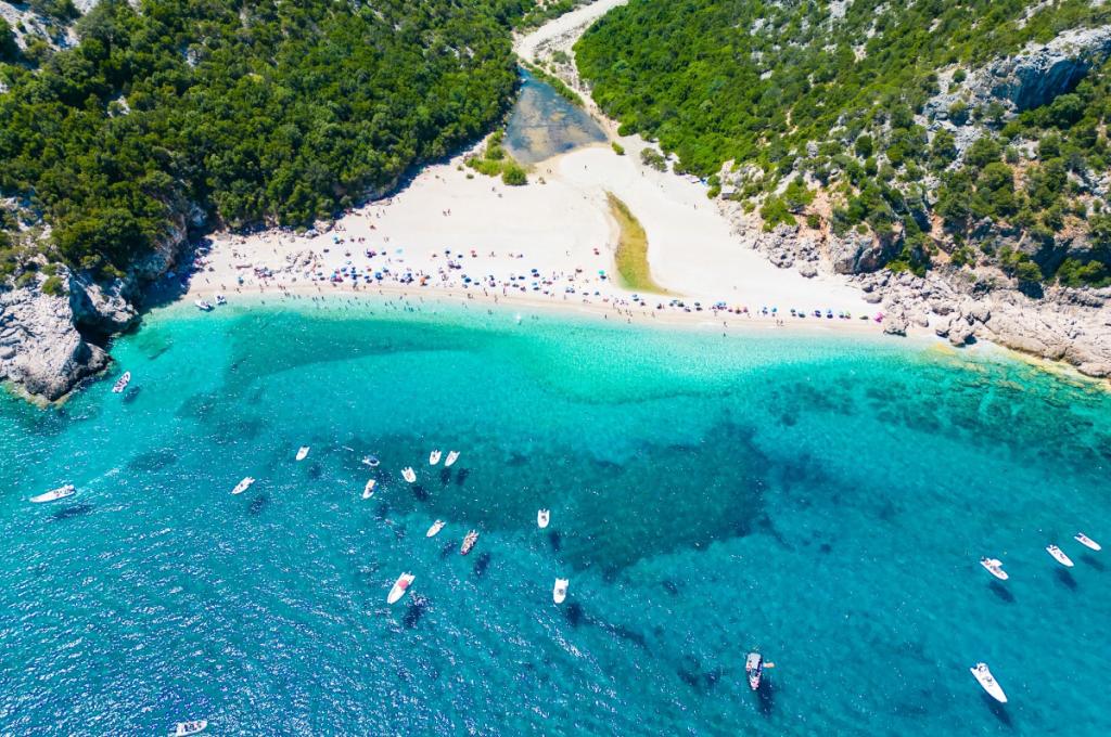 Spiaggia con acque turchesi, barche e ombrelloni, circondata da verde e rocce.