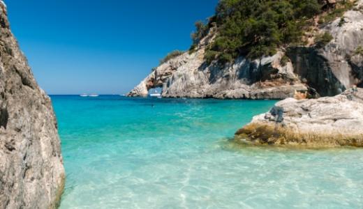 Spiaggia con acque cristalline, rocce e vegetazione lussureggiante sotto un cielo sereno.